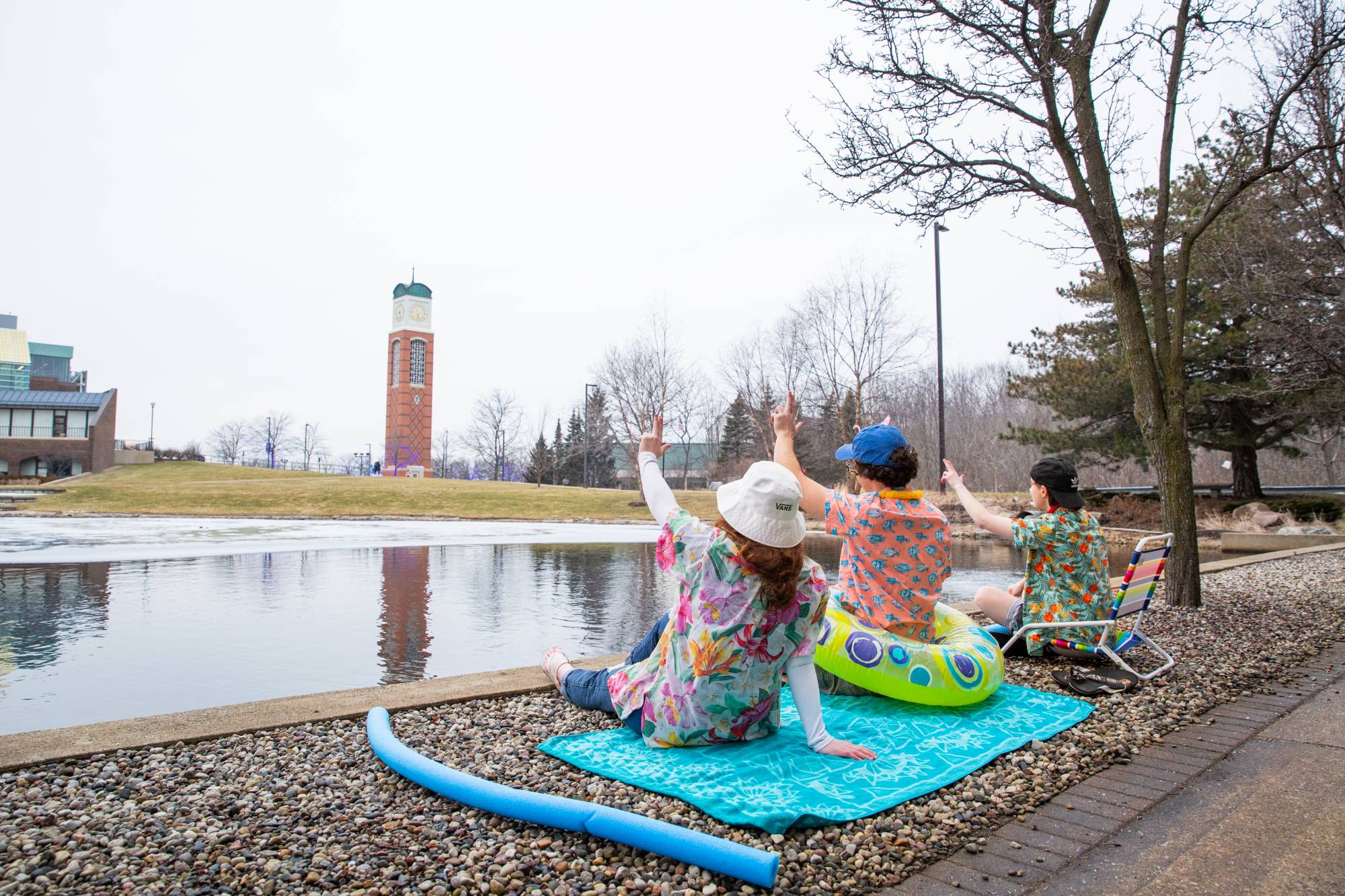 Three students dressed in beach attire sit on the ground behind Zumberge Pond. They're facing the clock tower and raising their hands, holding the "anchor up" hand signal.